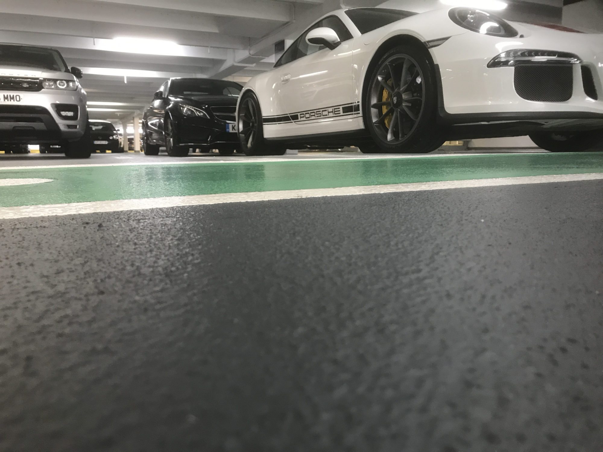 Low-angle view of several parked cars, including a white Porsche, in a multi-storey indoor car park with a green-striped floor.