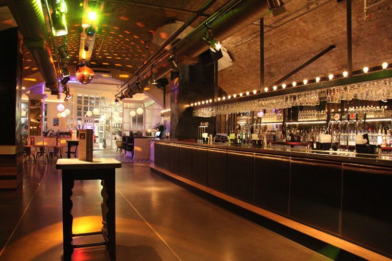 A modern bar interior with colourful lights, a long counter lined with glasses, empty tables, and industrial-style decor.