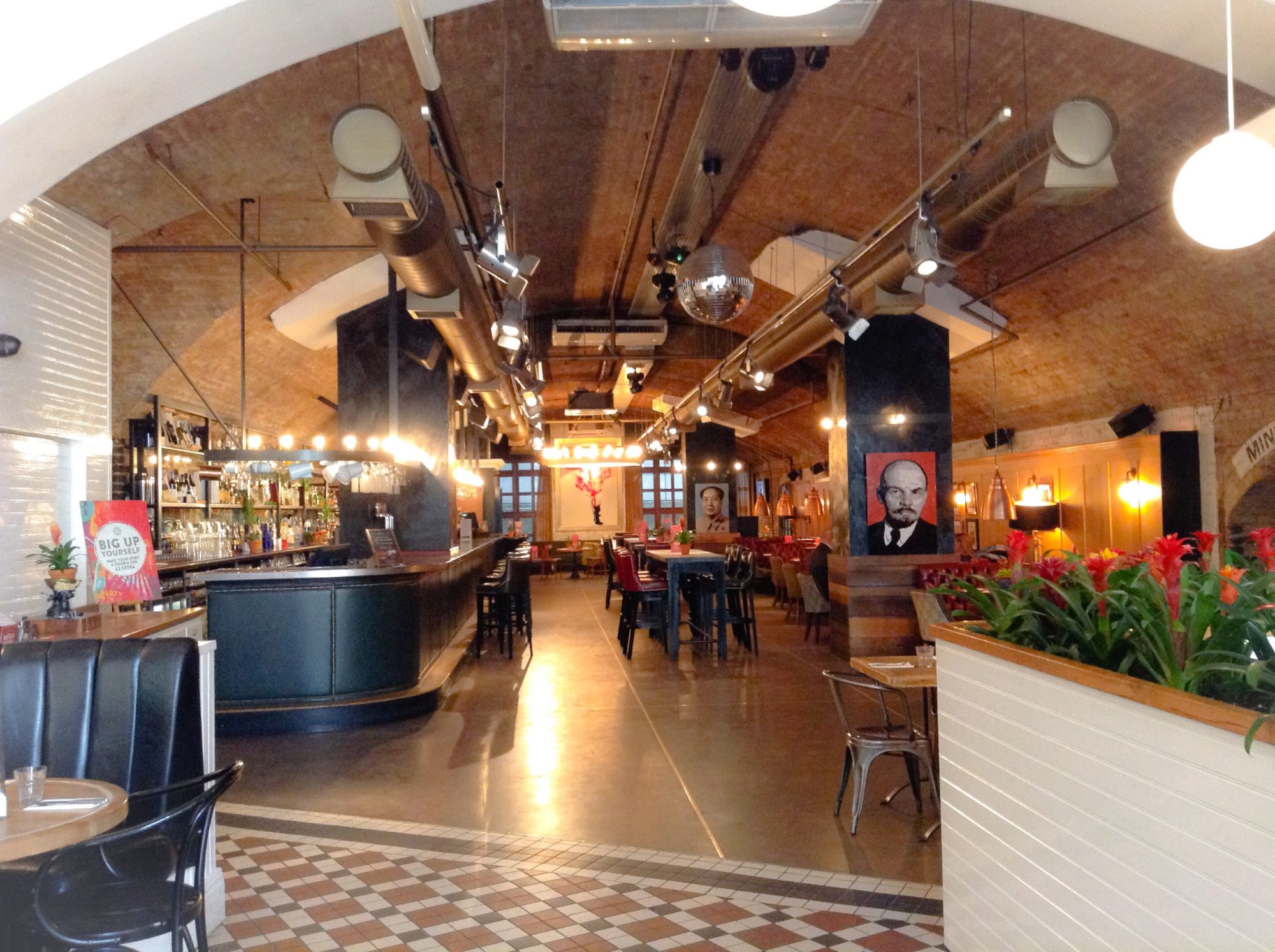 Restaurant with vaulted brick ceiling, modern lighting, bar on the left, tables in the centre, plant on the right, and portrait artwork on the far wall and column.