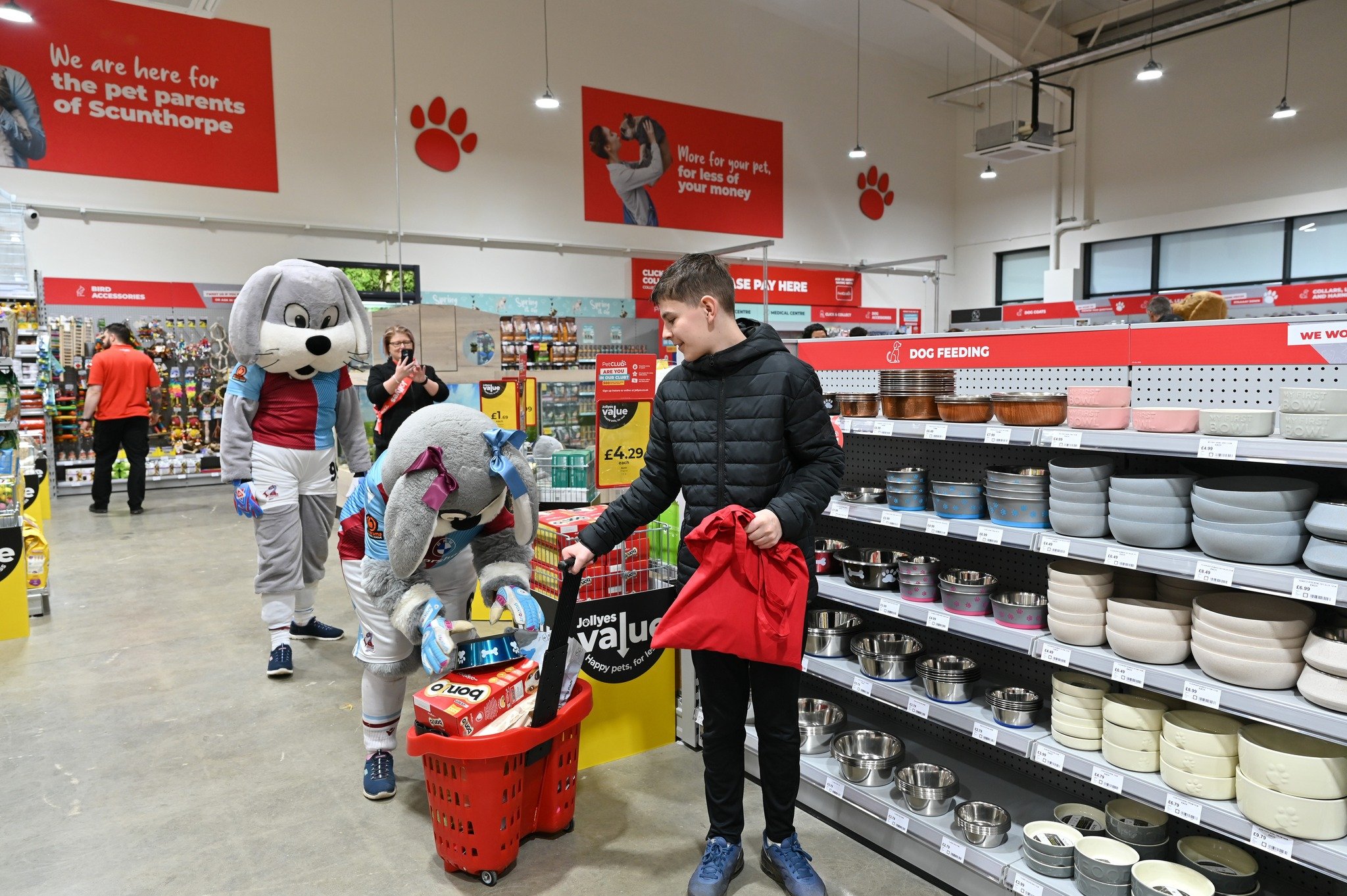 Two people in dog costumes and a boy shop in a pet shop aisle lined with bowls and pet supplies. The boy holds a red shirt and smiles as the mascot adds items to a red basket.